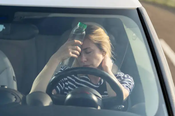 Exhausted woman driver feeling headache, sitting inside her car, applies bottle of water to forehead, hot weather. Tired female stop after driving car in traffic jam. Blood pressure, heat concept.