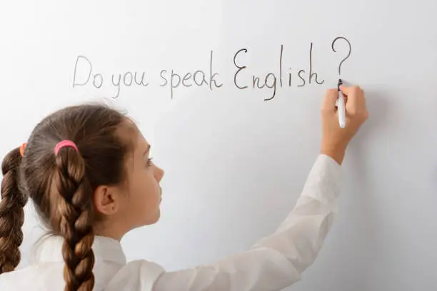 Small school girl writing Do you speak English? inscription on the blackboard