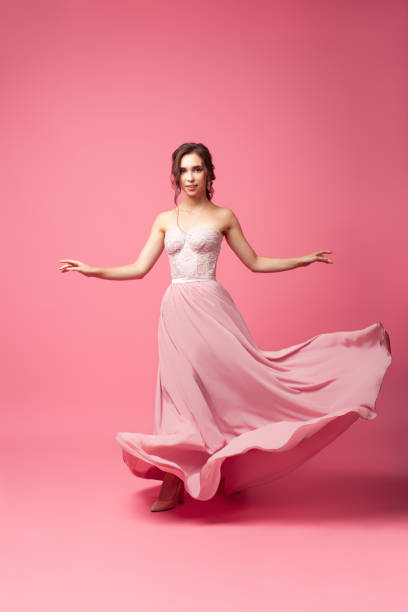 A young woman in a silk dress on a pink isoleted background. A model woman in a long evening dress is spinning in a dance A young woman in a silk dress on a pink isoleted background. A model woman in a long evening dress is spinning in a dance. skirt photos stock pictures, royalty-free photos & images