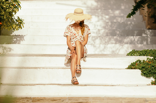 Gorgeous young woman sitting on white stairs outside and enjoying sunny day. She wears short dress and hat and looks very attractive. She is unrecognizable