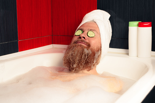 Lindo hombre barbudo tomando baño con la cabeza envuelta en toalla y rodajas de pepino en sus ojos. Divertido hipster relajándose en la bañera espumosa y disfrutando de la vida. SPA en casa. photo