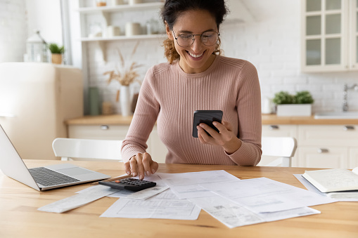 Smiling young 30s woman in eyewear looking at smartphone screen, feeling satisfied with fast secure online service, paying household bills taxes or insurance, managing budget, calculating expenses.