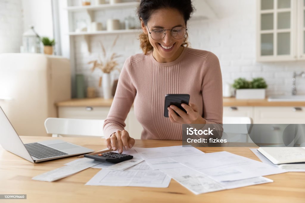 Smiling young woman satisfied with online payment mobile service. Smiling young 30s woman in eyewear looking at smartphone screen, feeling satisfied with fast secure online service, paying household bills taxes or insurance, managing budget, calculating expenses. Financial Bill Stock Photo
