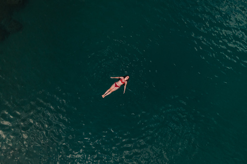 Young woman in red swimsuit floating on the sea surface, smiling and relaxing. Vacation time. Drone perspective. Aegean Sea