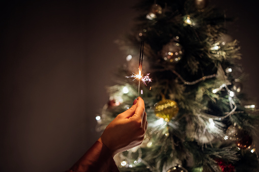 An anonymous woman holding a New Year's sparkler with a glowing decorated Christmas tree in the background