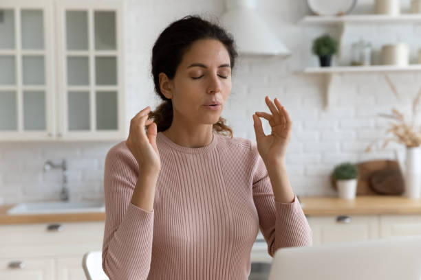 Mindful young woman calming down in stressful situation. Mindful young woman breathing out with closed eyes, calming down in stressful situation, working on computer in modern kitchen. Millennial hispanic lady managing stress, practice yoga at home office. inhaling stock pictures, royalty-free photos & images