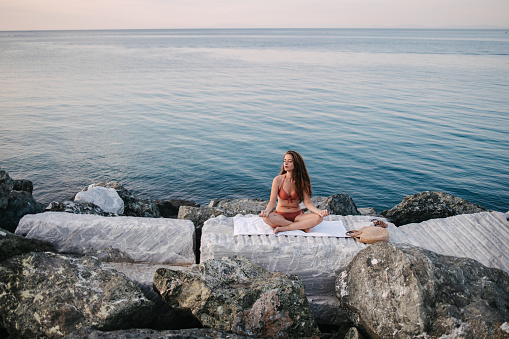 Woman meditating by the sea, sitting on the rocks
