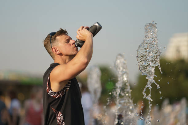 Handsome man drinking water in the hot summer day in the big city. Thermos with cold water. Fresh water splashes of fountain. Concept of leisure time, freshness and happiness. Portrait, frontal view