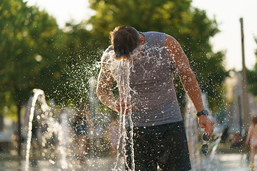 Handsome man washing his face by cold water of fountain in the hot summer day in the big city. Fresh water splashes. Concept of the leisure time, freshness and happiness. Frontal view