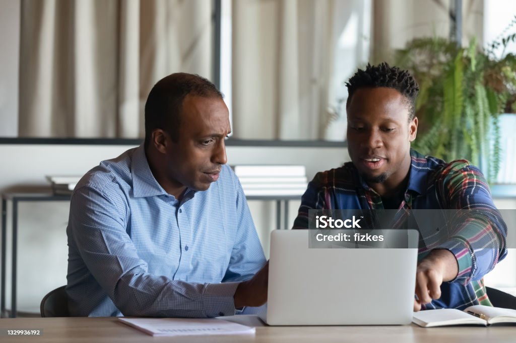 Two African American employees working on project together, using laptop Two African American employees working on project together, using laptop pointing at screen, discussing strategy and sharing ideas at meeting, colleagues involved in teamwork, cooperation concept Advice Stock Photo