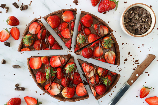 Stock photo showing an elevated view of a sliced, chocolate and strawberry tart with crispy, fluted, cocoa pastry crust, topped with whole and halved strawberries drizzled with melted chocolate.