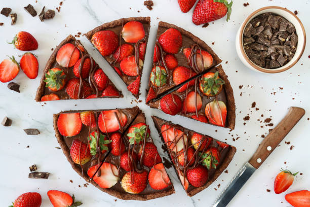 Image of sliced, chocolate strawberry tart, crispy, cocoa pastry crust, topped with whole and halved strawberries drizzled with melted chocolate, surrounded by strawberries, beside bowl of chocolate pieces, knife, marble effect background, elevated view stock photo
