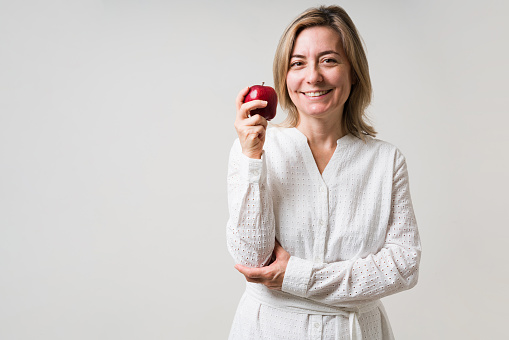 Starting a new nutritious diet. Healthy mature woman holding a delicious apple and ready to eat fruit against a gray studio background