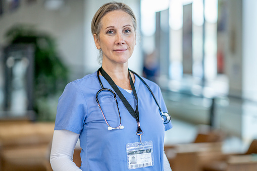 A nurse wearing blue scrubs smiles confidently at the camera. She is alone, but there are doctors and nurses walking around in the clinical space behind her.
