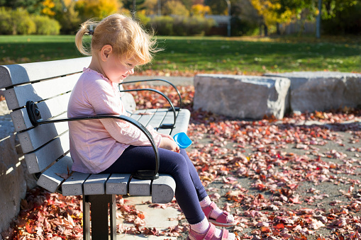 Child playing with leaves in autumn, little girl sitting on the bench during warm bright sunny day in the public local park