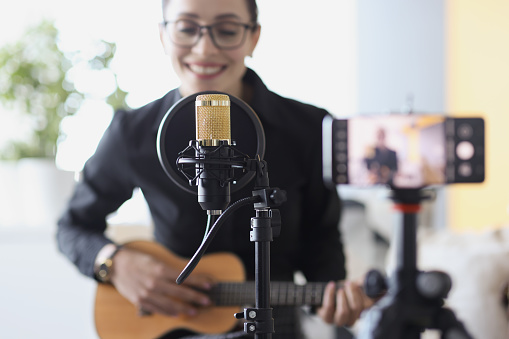 Woman plays guitar and sings while recording a video on smartphone. Guitar learning online concept