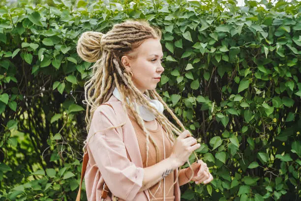 Woman with dreadlocks and white headphones on green plant background. Sunny summer day. Copy space.