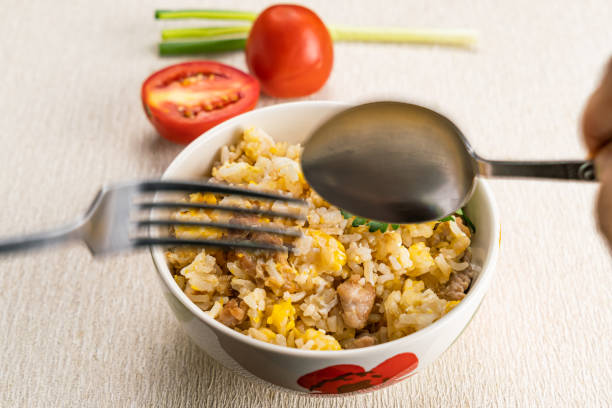 Man hand using metal spoon and fork scoop delicious thai style homemade pork and egg fried rice in ceramic bowl on table mat with whole and half tomato and green onion for his dinner.