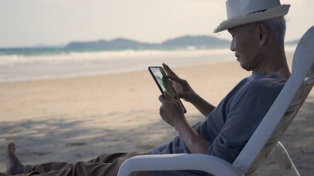 istock Senior man sitting on chairs enjoying time on beach travel vacation using tablet computer 1329990096