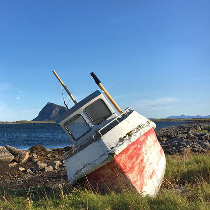 This old fishing boat is leaning sideways on a stony bank. The color has aged from the weather and the sun. There is a connection in the imposing landscape of Norway.