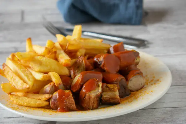 Homemade fresh cooked traditional german fast food meal with curry sausage and fries served with a delicious and spicy curry tomato sauce on a plate. Isolated and front view with wooden table background.
