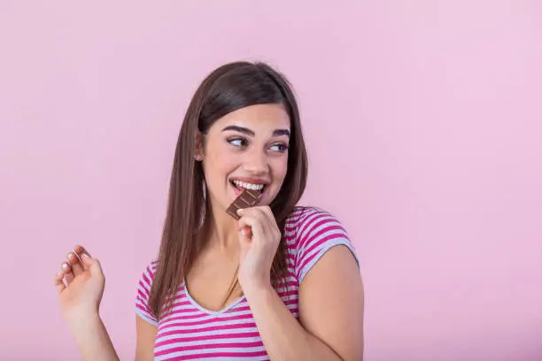 Photo of Happy young beautiful lady eating chocolate and smiling. Girl tasting sweet chocolate. Young woman with natural make up having fun and eating chocolate isolated on pink background