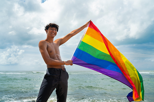 Portrait of young Hispanic gay boy looking at camera, holding LGBT flag - Focus on face
