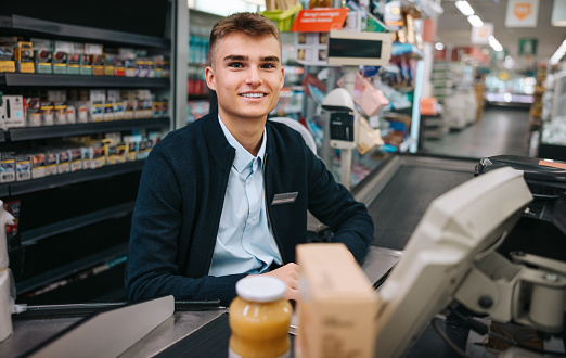 Male cash register working at grocery store checkout. Man sitting behind billing counter looking at camera and smiling.