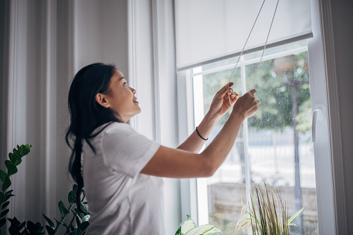 One woman, Asian female lowering interior blinds on window at home.