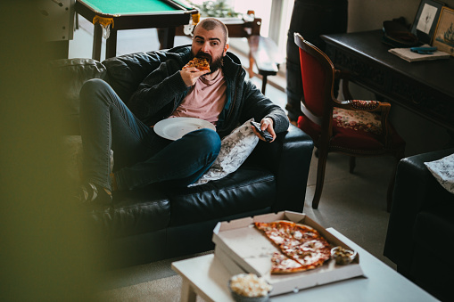 A young bearded man sitting at the couch in the living room, eating take-out pizza and watching TV