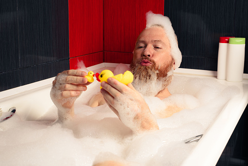Cute funny bearded man playing with rubber ducks in bubble bath relaxing at home.