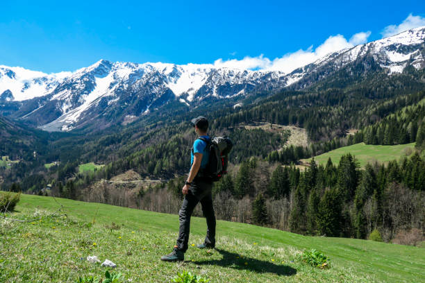 A man with a big hiking backpack enjoying the panoramic view on Baeren Valley in Austrian Alps. The highest peaks are snow-capped. Lush green pasture. Clear and sunny day. High mountain chains.