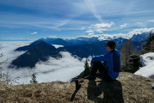A man in hiking outfit enjoying his morning tea with the panoramic view from the top of Alpine peak in Austria. The area is shrouded in thick clouds. A few peaks popping out from the clouds. Happy