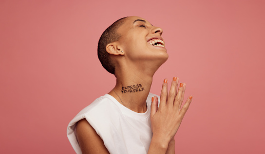Non binary female with shaved head smiling on pink background. Androgynous woman with express yourself written on neck.