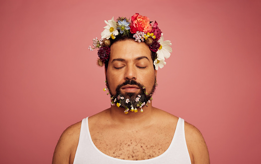 Gay man with flowers on head and beard. Gender fluid male with eyes closed on pink background.