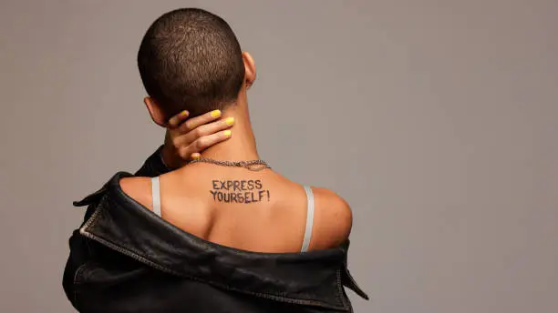 Rear view of anonymous woman with express yourself written on back. Gender fluid person from behind on grey background.