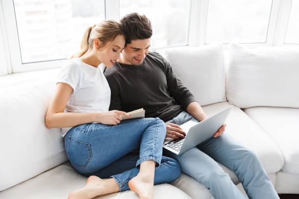 Image of happy couple using laptop and reading book while sitting on couch at home