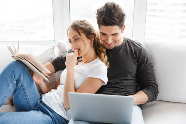 istock Image of modern couple using laptop and reading book while sitting on couch at home 1330034521