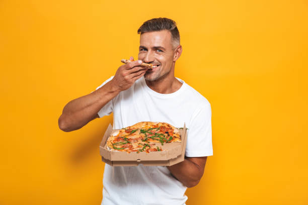 Image of joyful man 30s in white t-shirt holding and eating pizza while standing isolated Image of joyful man 30s in white t-shirt holding and eating pizza while standing isolated over yellow background isolated color stock pictures, royalty-free photos & images