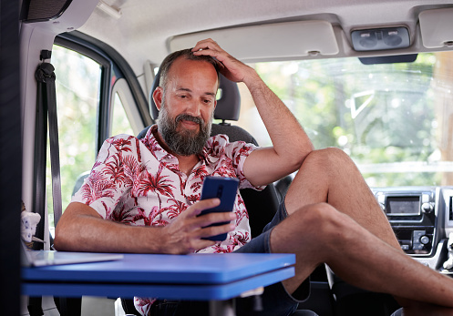 hombre barbudo de mediana edad con camisa de verano mirando sonrientemente su teléfono móvil dentro de su casa rodante photo