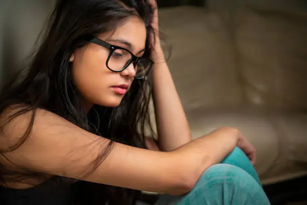 Photo of Sad, serene bespectacled young woman sitting near couch at night and contemplating.