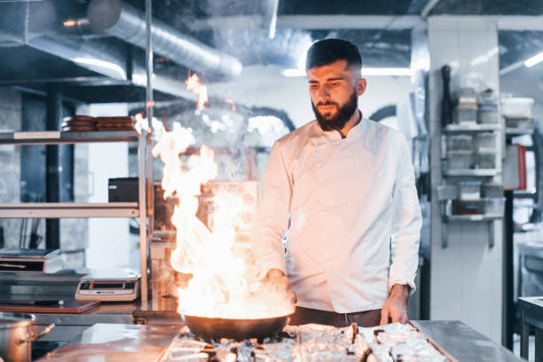 istock Frying pan is on fire. Chef in white uniform cooking food at kitchen. Busy day at work 1330044029