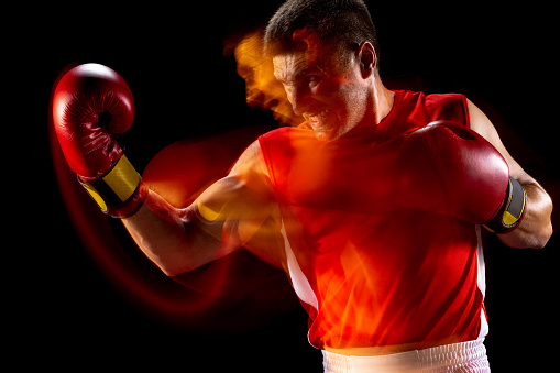 Punch, uppercut. Young sportsman, male boxer in red sportswear and gloves exercising on dark studio background in mixed lights. Copy space for ad.