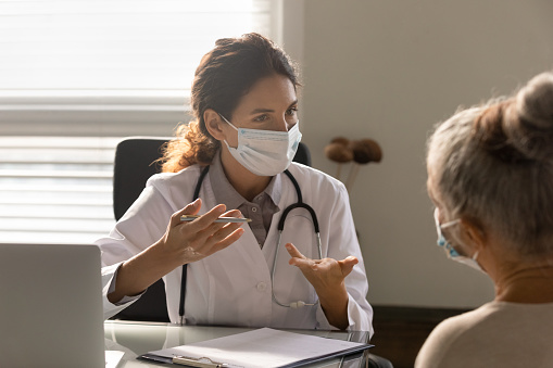 Young Caucasian female doctor in facial mask talk consult old patient in hospital or clinic. Woman GP in facemask against corona covid-19 have consultation with mature client. Coronavirus concept.