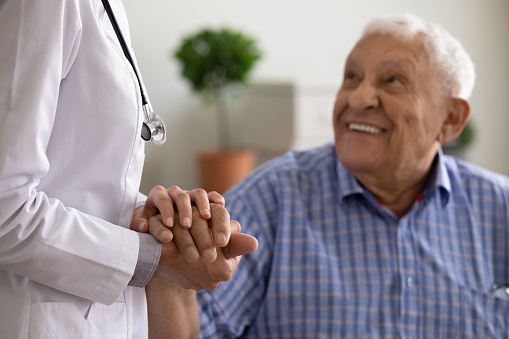 Close up of female nurse in white uniform hold happy mature male patient hand show good medical service. Woman doctor support comfort old man client in clinic or hospital. Elderly healthcare concept.