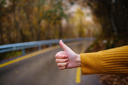 Close up shot of a womans hand doing hitchhiking sign near the country roadside during vacation trip in mountains fall forest