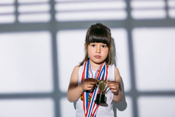 スポーツ選手権のメダルとトロフィーカップを持つ黒髪の女の子は、彼女の上にブラインドで窓の影に立っています。スポーツと勝利のコンセプト - humor athlete trophy one person ストックフォトと画像