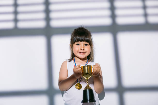 ragazza dai capelli scuri con medaglie del campionato sportivo e coppa dei trofei, in piedi all'ombra di una finestra con un cieco su di lei. concetto di sport e vittoria - humor athlete trophy one person foto e immagini stock