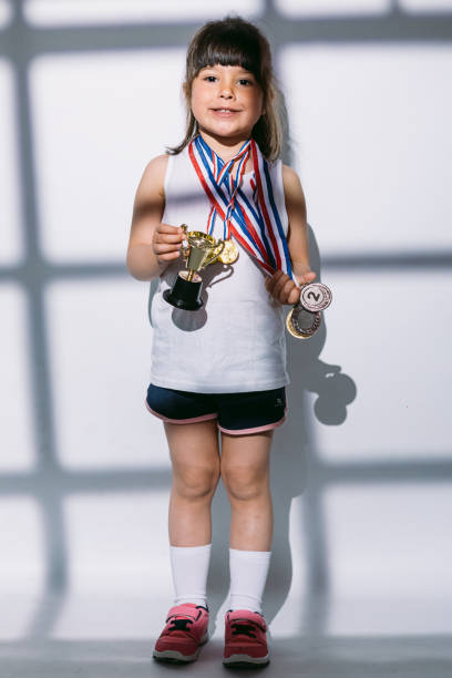 スポーツ選手権のメダルとトロフィーカップを持つ黒髪の女の子は、彼女の上にブラインドで窓の影に立っています。スポーツと勝利のコンセプト - humor athlete trophy one person ストックフォトと画像
