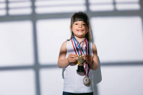 ragazza dai capelli scuri con medaglie del campionato sportivo e coppa dei trofei, in piedi all'ombra di una finestra con un cieco su di lei. concetto di sport e vittoria - humor athlete trophy one person foto e immagini stock